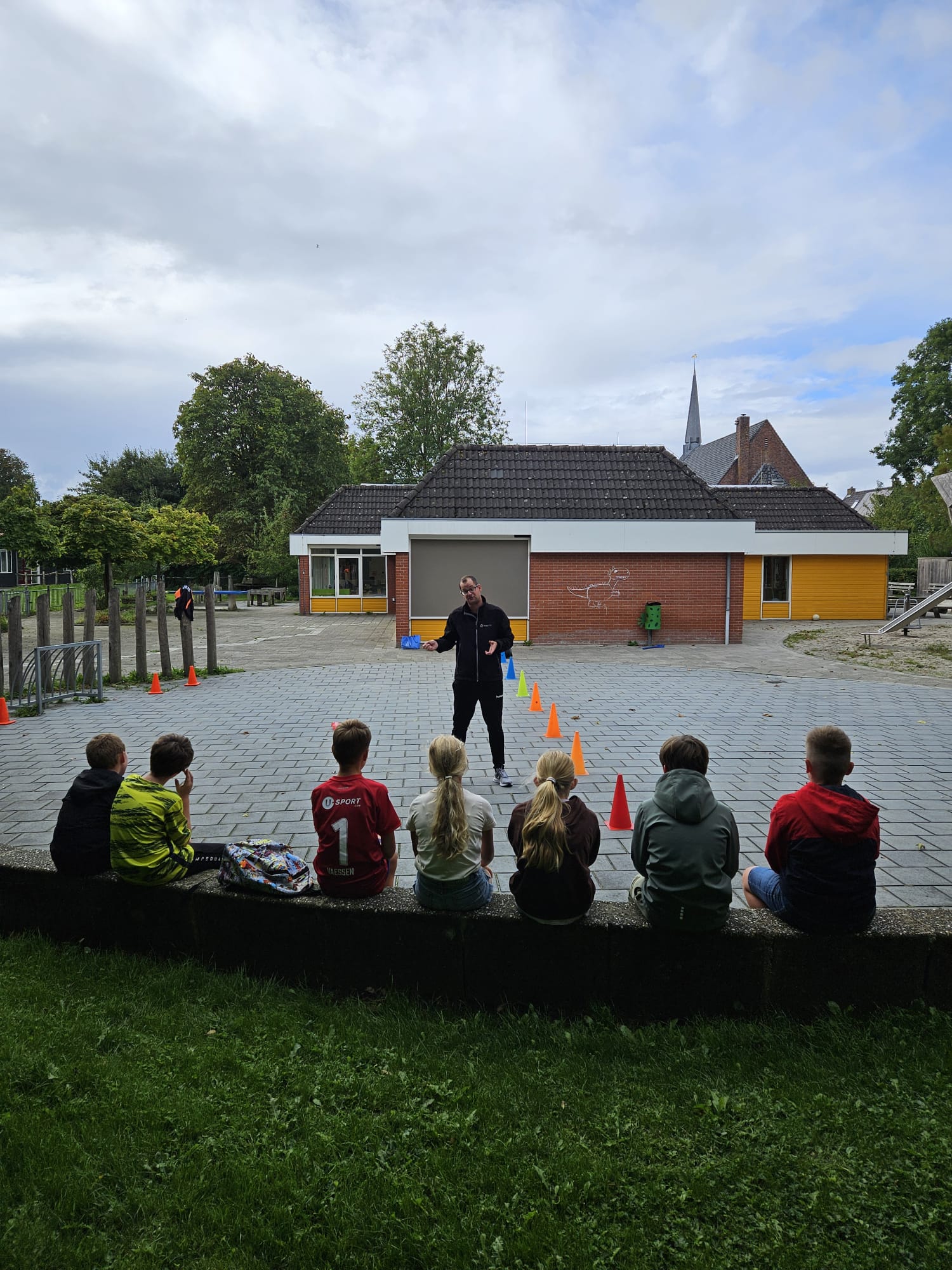Kinderen zitten op een muurtje bij het schoolplein van ODBS Abt Emo en luisteren naar de uitleg van de sportdocent